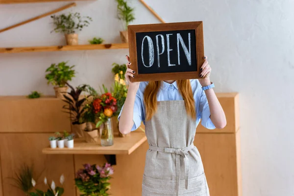 Young female florist in apron hiding face behind open sign in flower shop — Stock Photo