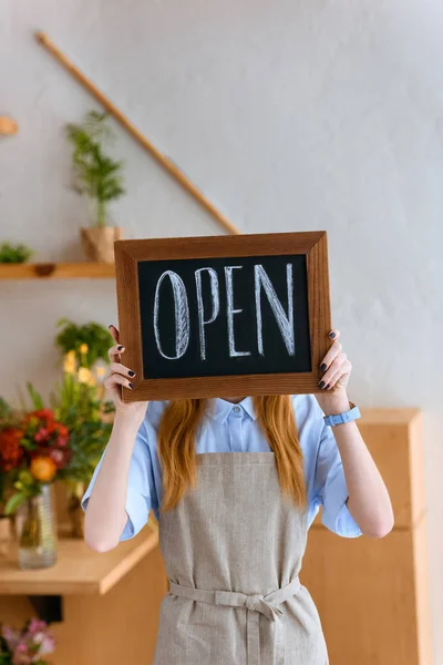 Junge Floristin in Schürze versteckt Gesicht hinter offenem Schild in Blumenladen — Stockfoto