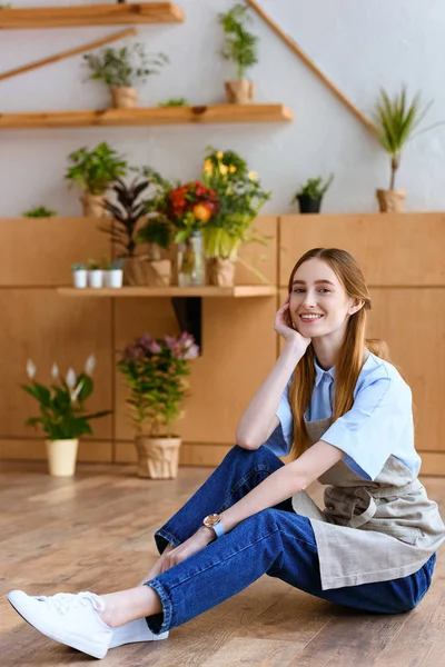 Hermosa florista joven sentado en el suelo y sonriendo a la cámara en la tienda de flores - foto de stock