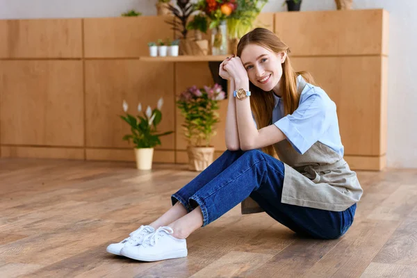 Beautiful young florist sitting on floor and smiling at camera in flower shop — Stock Photo