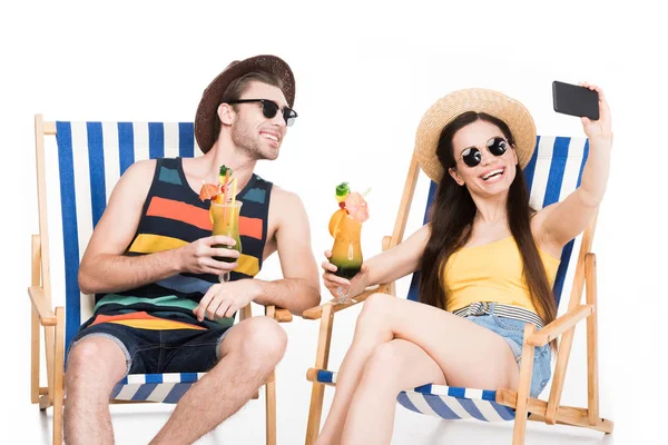 Couple souriant reposant sur des chaises de plage avec des cocktails et de prendre selfie, isolé sur blanc — Stock Photo