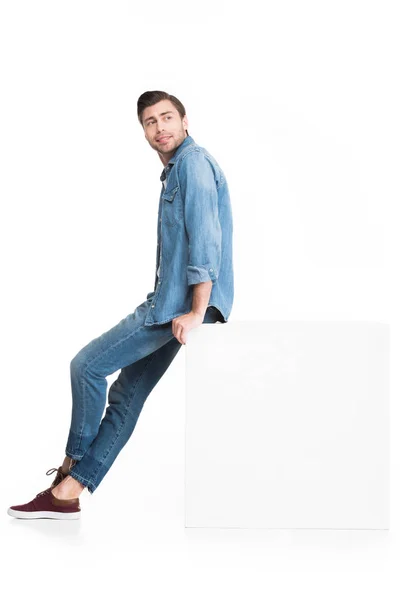 Young smiling man in jeans posing at white cube, isolated on white — Stock Photo