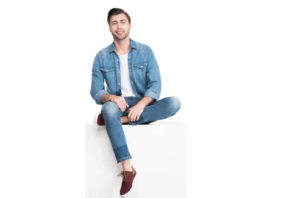 Молодий усміхнений чоловік в джинсах сидить на білому кубі, ізольований на білому — стокове фото