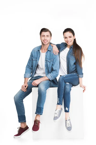 Coppia sorridente in jeans seduta su cubo bianco e guardando la macchina fotografica, isolata su bianco — Foto stock