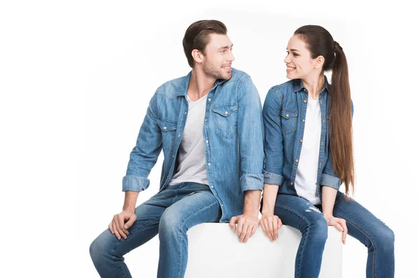 Cheerful couple sitting on white cube and looking at each other, isolated on white — Stock Photo