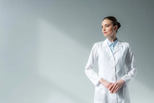 Séduisante femme médecin en manteau blanc regardant loin sur gris — Photo de stock