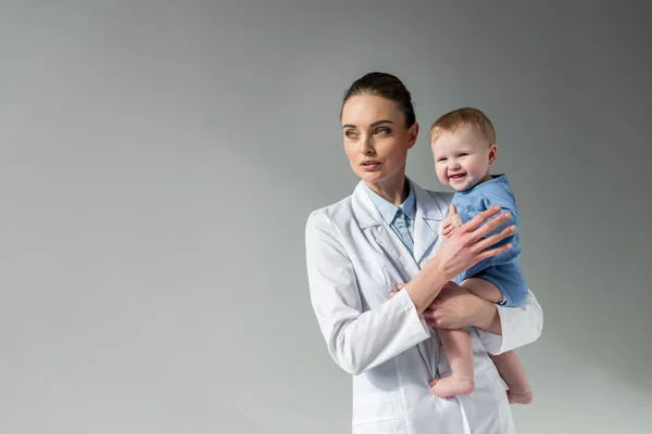 Hermosa mujer pediatra sosteniendo riendo riendo pequeño bebé en gris — Stock Photo