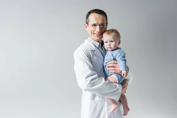 Handsome adult pediatrician holding little baby and looking at camera isolated on grey — Stock Photo