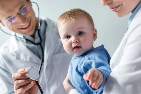 Recortado tiro de pediatras chequeando aliento de adorable pequeño bebé - foto de stock