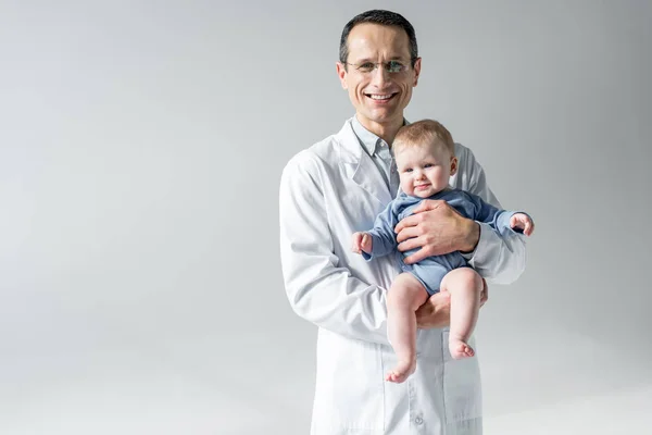 Glücklicher erwachsener Kinderarzt mit kleinem Baby und Blick in die Kamera isoliert auf grau — Stockfoto