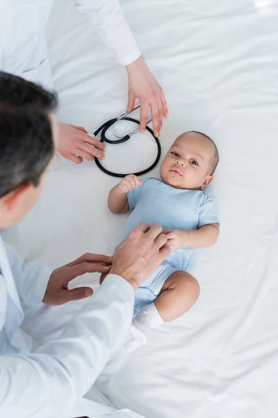 Recortado tiro de pediatras cosquillas pequeño bebé en la cama - foto de stock