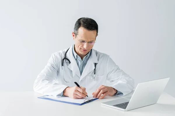 Serious adult doctor sitting at workplace and writing diagnosis in clipboard isolated on white — Stock Photo