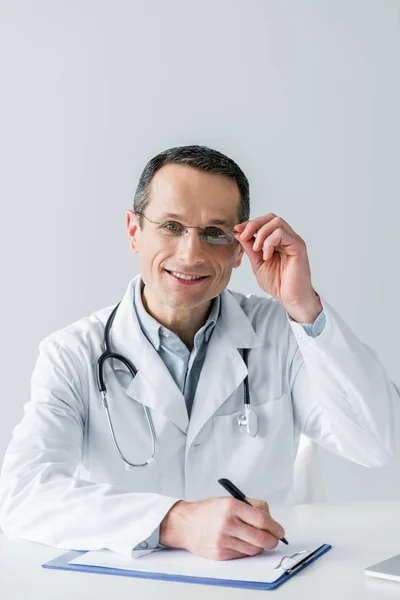 Médico adulto feliz sentado en el lugar de trabajo y mirando a la cámara mientras escribe en portapapeles aislado en blanco - foto de stock