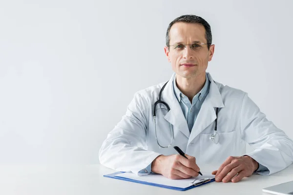 Guapo médico adulto sentado en el lugar de trabajo y escribir diagnóstico en portapapeles aislado en blanco - foto de stock