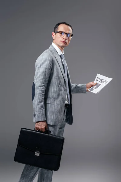 Hombre de negocios adulto guapo con maletín y periódico mirando a la cámara en gris - foto de stock