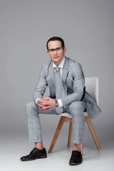 Homme d'affaires adulte souriant en costume élégant assis sur une chaise et regardant la caméra sur le gris — Photo de stock