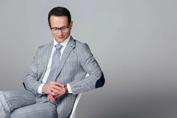 Handsome adult businessman in suit sitting on chair and looking at watch isolated on grey — Stock Photo