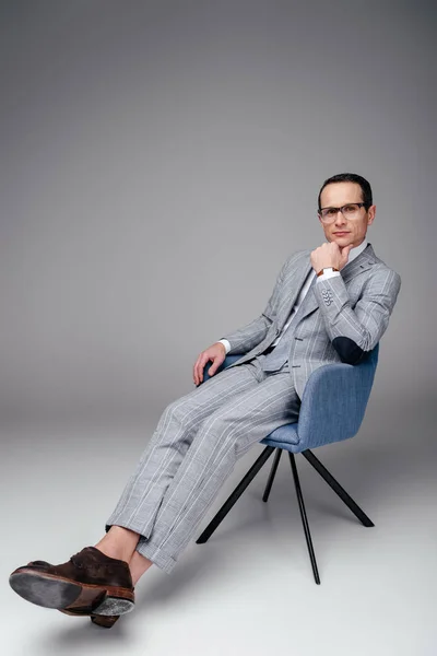 Adulto hombre de negocios en traje elegante sentado en silla con la mano en la barbilla en gris - foto de stock