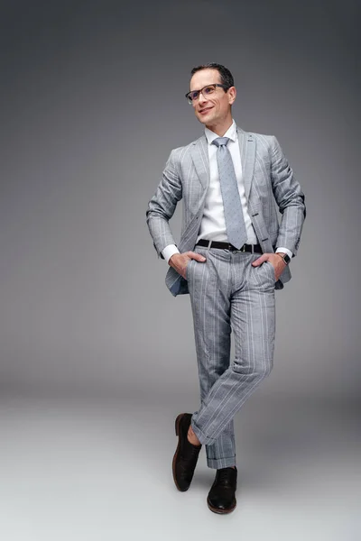 Guapo hombre de negocios maduro en traje elegante con manos en bolsillos en gris - foto de stock