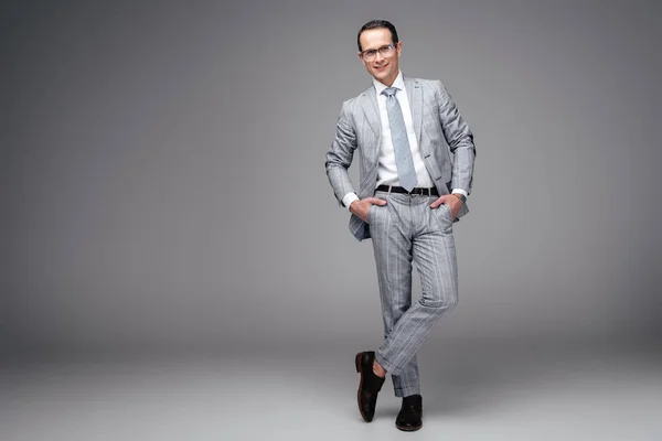 Красивий дорослий бізнесмен у стильному костюмі з руками в кишенях на сірому — стокове фото