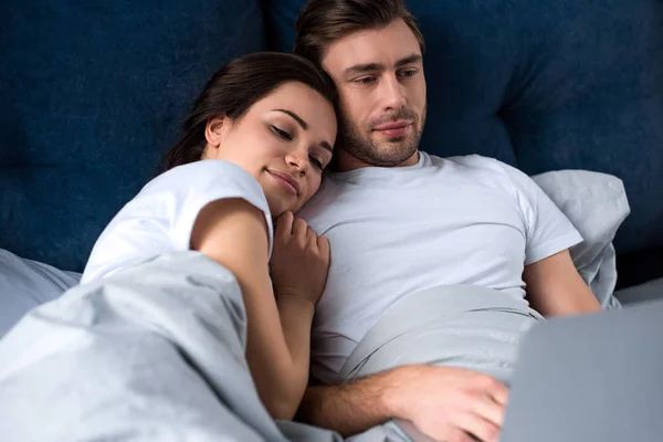 Усміхнена пара дивиться на ноутбук, лежачи в ліжку — стокове фото