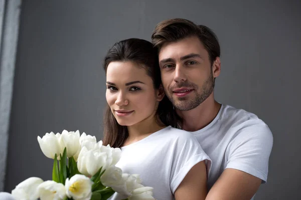 Улыбающиеся мужчина и женщина с цветами нежно обнимаются в спальне — стоковое фото