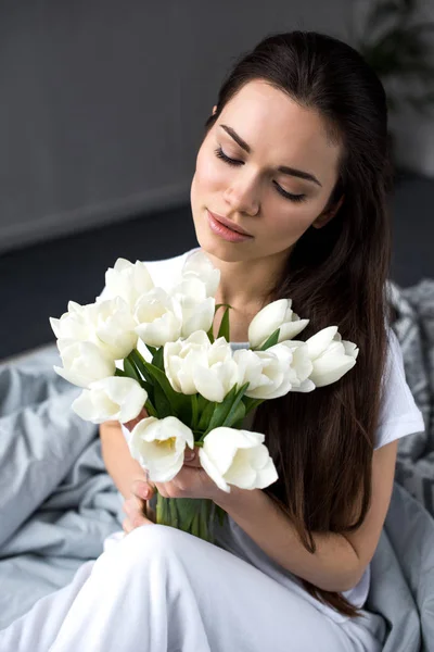 Dreamy girl sitting on bed and holding bouquet of tulips — Stock Photo