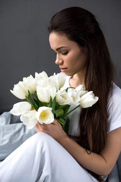 Attractive woman sitting on bed and holding bouquet of tulips — Stock Photo