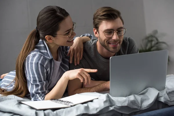 Joven pareja atractiva con bloc de notas mirando a la computadora portátil mientras está acostado en la cama - foto de stock