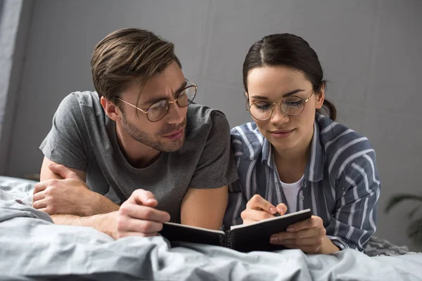 Nachdenklicher Mann schaut Frau beim Schreiben im Notizblock an, während sie im Bett liegt — Stockfoto