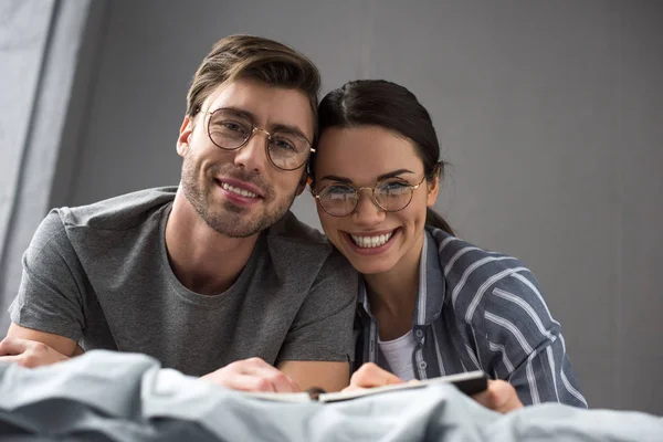 Улыбающаяся пара в очках пишет в блокноте, лежа в постели — стоковое фото
