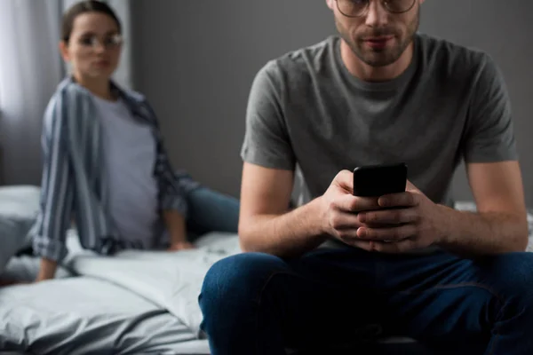 Женщина смотрит на мужчину, сидящего на кровати и использующего смартфон — стоковое фото