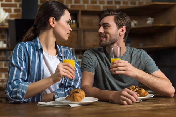 Smiling man and woman drinking juice and eating croissants by kitchen table — Stock Photo
