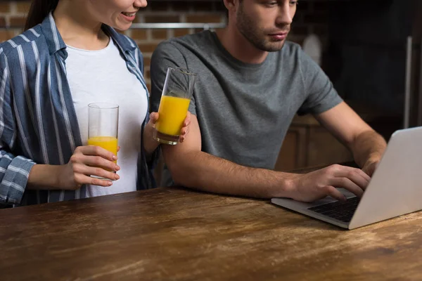 Mujer joven sosteniendo vasos con jugo mientras el hombre trabaja en el ordenador portátil - foto de stock
