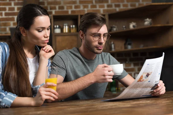 Mujer joven bebiendo jugo mientras el hombre toma café y lee el periódico - foto de stock