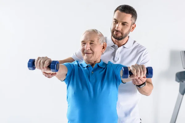 Retrato del terapeuta de rehabilitación que ayuda al hombre mayor a hacer ejercicio con pesas sobre fondo gris - foto de stock