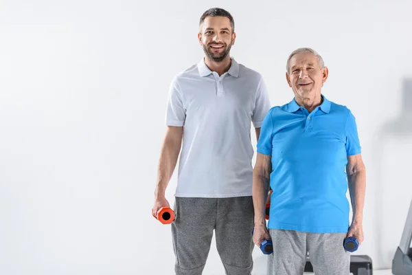 Smiling rehabilitation therapist and senior man with dumbbells looking at camera on grey background — Stock Photo