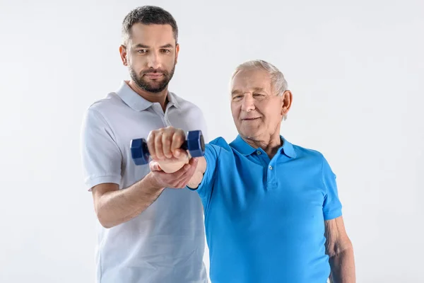 Retrato del terapeuta de rehabilitación que ayuda al hombre mayor a hacer ejercicio con mancuerna aislada en gris - foto de stock