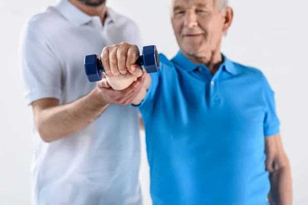 Visão parcial do terapeuta de reabilitação auxiliando o homem idoso a se exercitar com halteres isolados em cinza — Fotografia de Stock