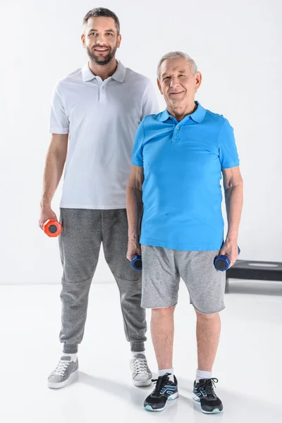 Terapeuta de reabilitação e homem idoso com halteres olhando para a câmera em fundo cinza — Fotografia de Stock