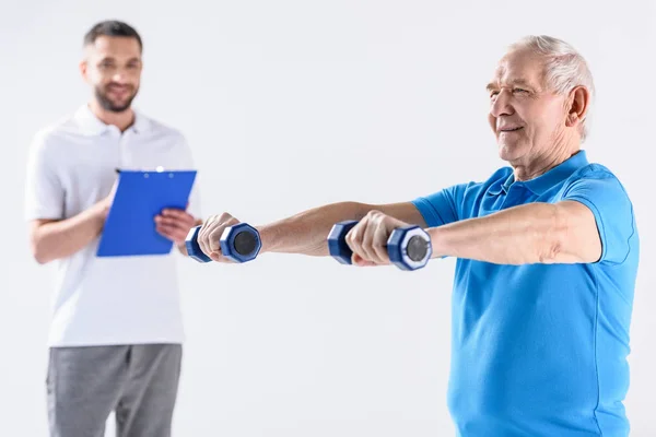 Retrato del terapeuta rehabilitador con bloc de notas asistiendo a un hombre mayor que hace ejercicio con pesas aisladas en gris - foto de stock