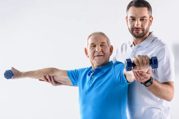 Portrait d'un thérapeute en réadaptation aidant un aîné à faire de l'exercice avec des haltères sur fond gris — Photo de stock
