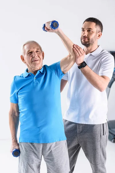 Retrato del terapeuta de rehabilitación que ayuda al hombre mayor a hacer ejercicio con pesas sobre fondo gris - foto de stock