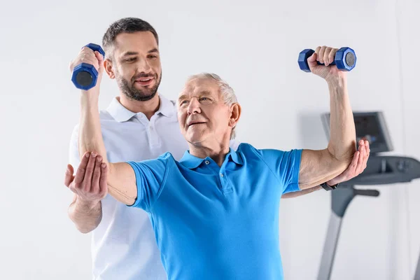 Retrato del terapeuta de rehabilitación que ayuda al hombre mayor a hacer ejercicio con pesas sobre fondo gris - foto de stock