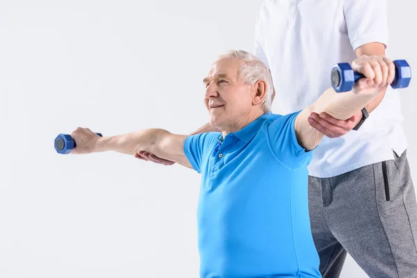 Vista parziale del terapeuta riabilitativo che assiste l'uomo anziano che si esercita con i manubri su sfondo grigio — Foto stock