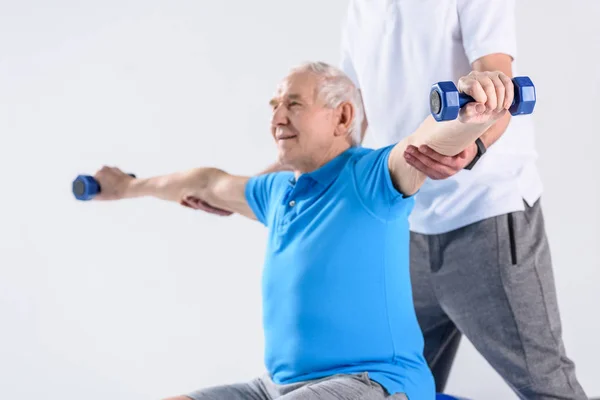 Tiro recortado de terapeuta de rehabilitación que ayuda a un hombre mayor a hacer ejercicio con pesas sobre fondo gris - foto de stock