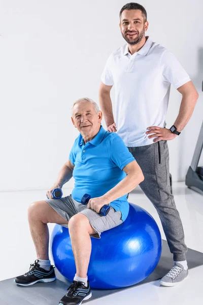 Sonriente terapeuta de rehabilitación akimbo y hombre mayor con mancuernas en la pelota de fitness - foto de stock