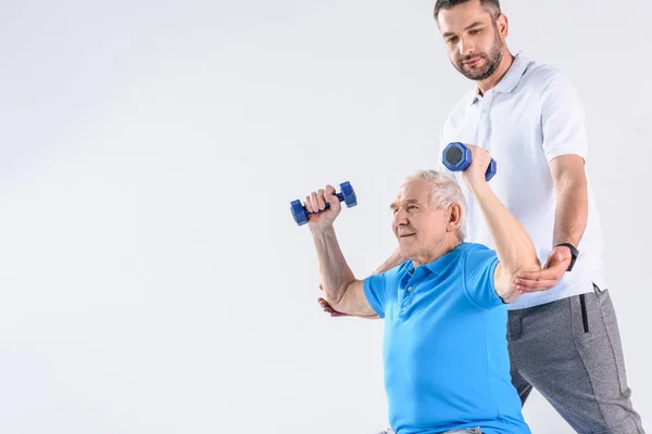 Retrato del terapeuta de rehabilitación que ayuda al hombre mayor a hacer ejercicio con pesas sobre fondo gris - foto de stock