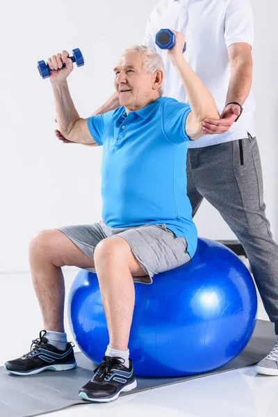 Plan recadré d'un thérapeute de réadaptation aidant un aîné à faire de l'exercice avec des haltères sur une balle de fitness isolée sur du gris — Photo de stock