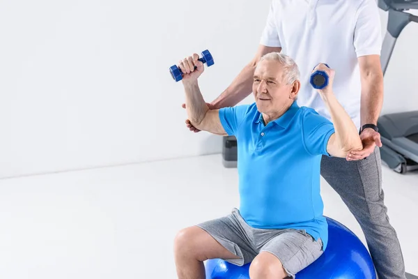 Vista parcial del terapeuta de rehabilitación que ayuda a un hombre mayor a hacer ejercicio con pesas sobre una pelota de fitness sobre un fondo gris - foto de stock